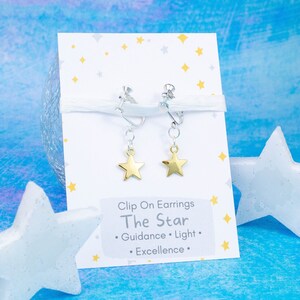 Peut inclure: Une paire de boucles d'oreilles clips en forme d'étoile dorée sur une carte blanche avec le texte "Clip On Earrings The Star  • Guidance • Light • Excellence •".