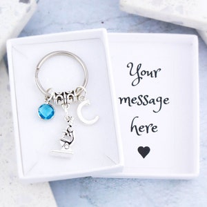 May include: A silver keychain with a microscope charm, a blue crystal, a letter C charm, and a silver ring. The keychain is in a white gift box with the text "Your message here" and a black heart.