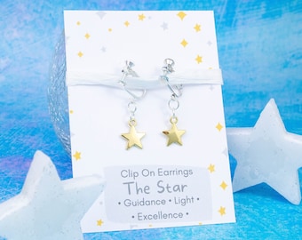 May include: A pair of gold star clip-on earrings on a white card with the text "Clip On Earrings The Star  • Guidance • Light • Excellence •".