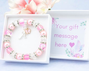 Puede incluir: Un brazalete de cuentas rosa y blanco con un dije de plata en forma de mariposa y la letra A. El brazalete está en una caja de regalo blanca con un diseño floral rosa y el texto "Tu mensaje de regalo aquí".