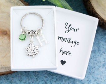 May include: A silver keychain with a green crystal charm, a silver maple leaf charm, and a silver letter "H" charm. The keychain is in a white box with the text "Your message here" and a heart.