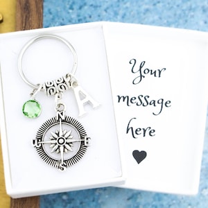 May include: A silver compass keychain with a green crystal charm and a silver letter A charm. The keychain is in a white box with the text "Your message here" and a small heart.