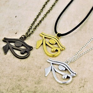 May include: Three necklaces with the Eye of Horus pendant. The pendants are made of different metals: bronze, gold, and silver. The necklaces are on a light brown surface.