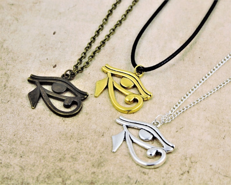 May include: Three necklaces with the Eye of Horus pendant. The pendants are made of different metals: bronze, gold, and silver. The necklaces are on a light brown surface.