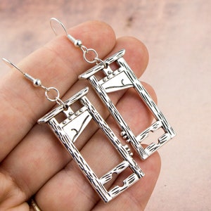 Puede incluir: Pendientes de guillotina de plata. Los pendientes tienen forma de guillotina con una cuchilla y un marco de madera. Los pendientes cuelgan de ganchos de plata.