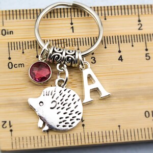 May include: Silver keychain with a pink crystal charm, a silver hedgehog charm, and a silver letter A charm. The keychain is measured against a wooden ruler.