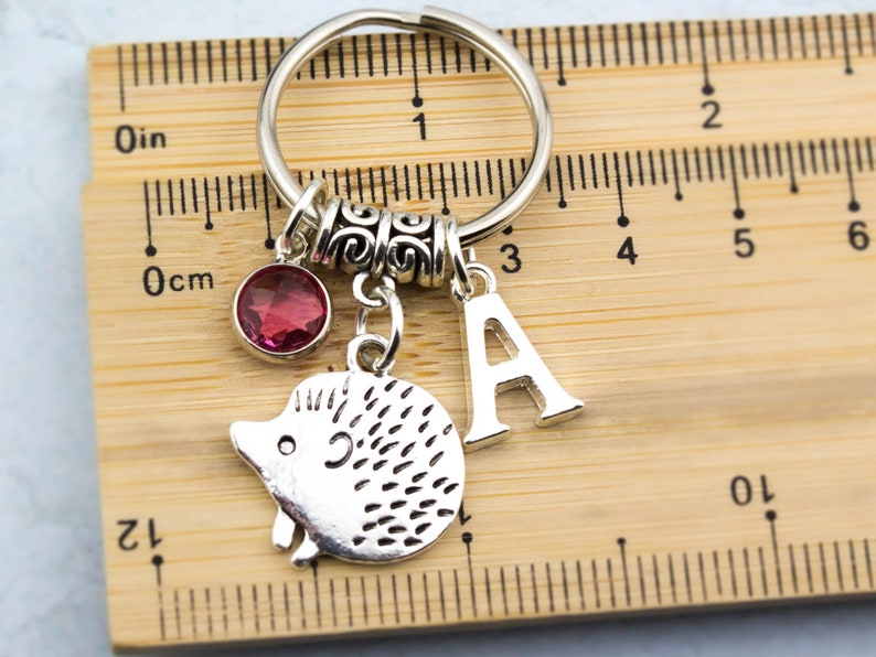 May include: Silver keychain with a pink crystal charm, a silver hedgehog charm, and a silver letter A charm. The keychain is measured against a wooden ruler.