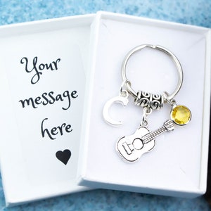 May include: A silver keychain with a guitar charm, a letter C charm, a small yellow crystal charm, and a silver ring. The keychain is in a white gift box with the text "Your message here" and a heart.