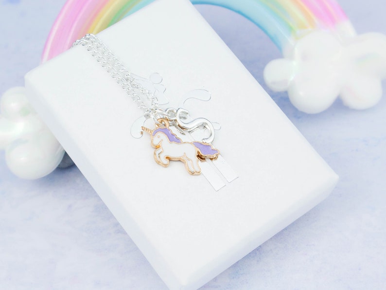 Op de afbeelding: Een zilveren ketting met een paarse en witte eenhoornbedel en een zilveren bedel. De ketting wordt gepresenteerd op een witte doos met een regenboogachtergrond.