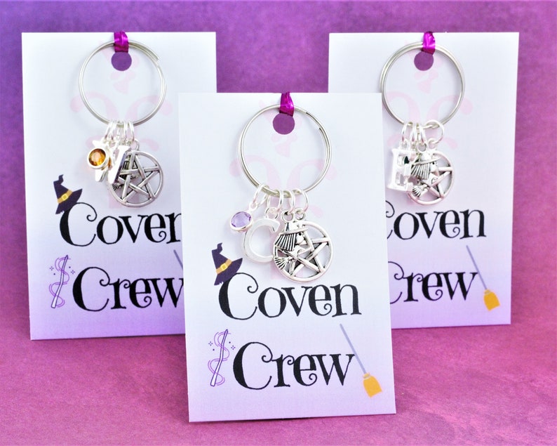 Puede incluir: Tres llaveros de plata con dijes, cada uno con un pentagrama, un sombrero de bruja y una escoba. Los llaveros se muestran en tarjetas blancas con el texto "Coven Crew" impreso en ellos.