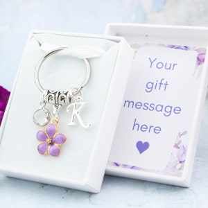May include: A silver keychain with a purple flower charm, a clear crystal, and the letter "K" in a white gift box. The box contains a card with the text "Your gift message here" and a purple heart.