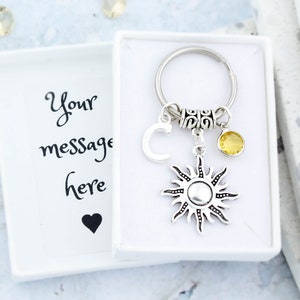 May include: A silver sun charm keyring with a yellow crystal charm and a silver letter C charm. The keyring is in a white box with the text "Your message here" and a black heart.