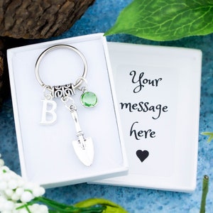 Puede incluir: Un llavero plateado con un pequeño encanto de pala, una piedra preciosa verde y un encanto de letra 'B'. El llavero está en una caja blanca con una tarjeta en blanco que dice 'Tu mensaje aquí'.