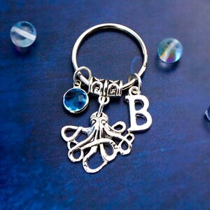 May include: Silver octopus keychain with a blue crystal charm and a silver letter 'B' charm. The keychain is on a blue background.