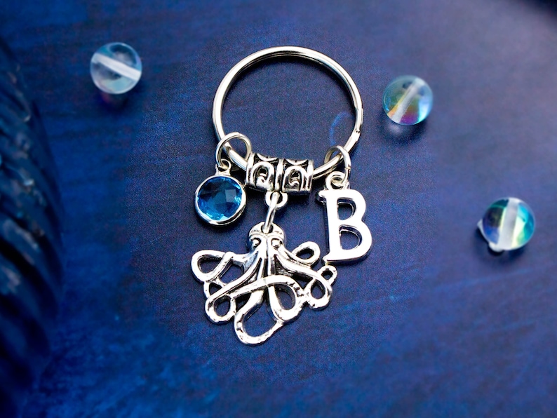 May include: Silver octopus keychain with a blue crystal charm and a silver letter 'B' charm. The keychain is on a blue background.