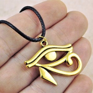 May include: A gold-colored Eye of Horus pendant necklace with a black braided cord. The pendant is a detailed representation of the ancient Egyptian symbol.