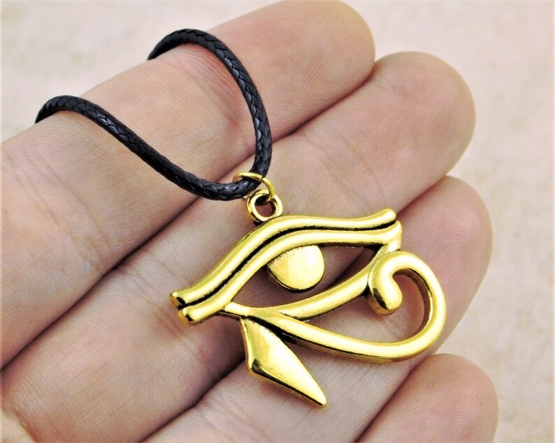 May include: A gold-colored Eye of Horus pendant necklace with a black braided cord. The pendant is a detailed representation of the ancient Egyptian symbol.