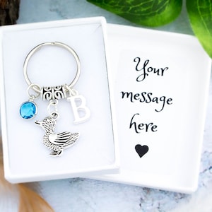 May include: A silver duck keyring with a blue crystal charm, a silver letter 'B' charm, and a silver ring. The keyring is in a white gift box with the text 'Your message here' and a black heart.