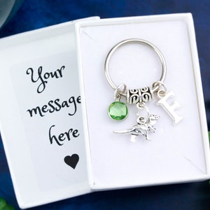 May include: A silver keyring with a green gem, a dinosaur charm, and a letter "F" charm. The keyring is in a white box with the text "Your message here" and a heart.