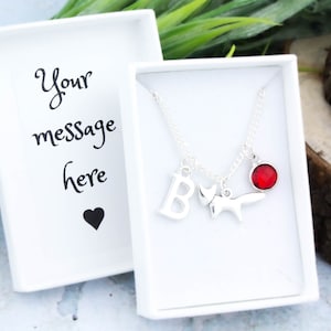 May include: A silver necklace with a fox charm, a letter B charm, and a red gemstone charm. The necklace is in a white gift box with the text "Your message here" and a heart.