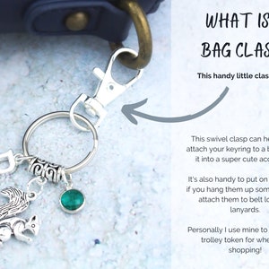 May include: A silver swivel clasp keychain with a green gem, a squirrel charm, and a letter "P" charm. The keychain is attached to a blue bag. The text "WHAT IS A BAG CLASP?" is written in white on a gray background. The text "This handy little clasp here!" is written below the title. The text below describes how to use the clasp to attach a keyring to a bag, hang keys, or attach to belt loops or lanyards.