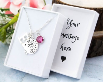 Puede incluir: Un collar de plata con un dije de erizo, un dije de corona y un dije de cristal rosa. El collar está en una caja de regalo blanca con el texto "Tu mensaje aquí" y un corazón.