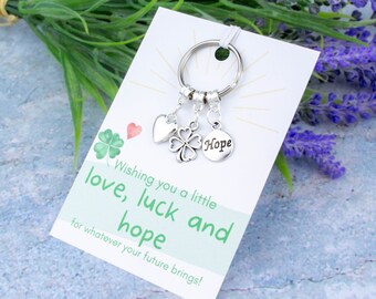 May include: A silver keychain with a heart, four-leaf clover, and a round charm with the word "Hope" on it. The keychain is attached to a white card with the text "Wishing you a little love, luck and hope for whatever your future brings!"