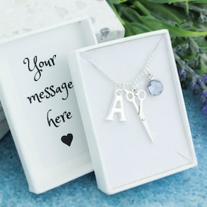 May include: A silver necklace with a letter A charm, a pair of scissors charm, and a light blue crystal charm. The necklace is in a white box with a blank card that says "Your message here" and a black heart.