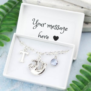 May include: A silver charm bracelet featuring a sloth charm, a letter T charm, and a light blue crystal charm. The bracelet is presented in a white gift box with the message "Your message here" and a heart.