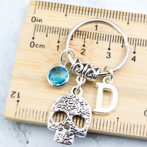 May include: Silver keychain with a skull charm, a blue crystal charm, a silver letter D charm, and a silver ring. The keychain is measured against a ruler.