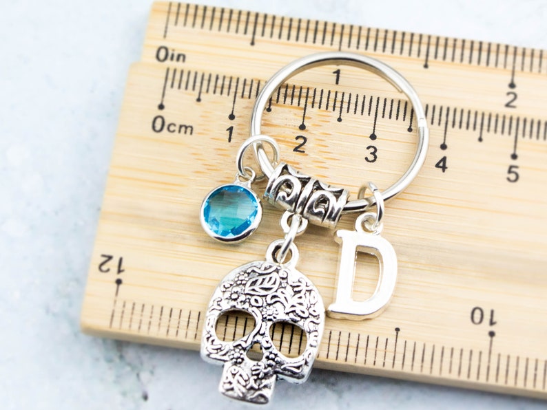 May include: Silver keychain with a skull charm, a blue crystal charm, a silver letter D charm, and a silver ring. The keychain is measured against a ruler.