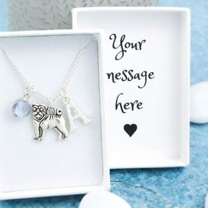 May include: A silver necklace with a charm of a pug, a letter A charm, and a light blue gemstone charm. The necklace is in a white box with the text "Your message here" and a black heart.
