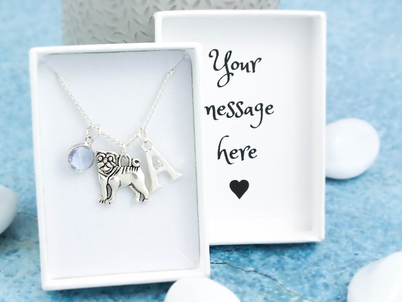 May include: A silver necklace with a charm of a pug, a letter A charm, and a light blue gemstone charm. The necklace is in a white box with the text "Your message here" and a black heart.