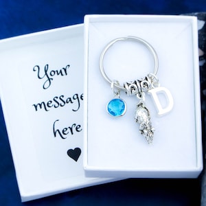 Puede incluir: Un llavero plateado con un dije de cristal azul, un dije de animal plateado y un dije de letra D plateado. El llavero est&aacute; en una caja blanca con una tarjeta en blanco que dice "Tu mensaje aqu&iacute;".