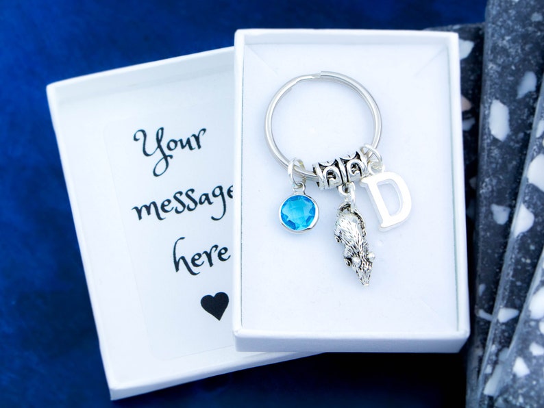 Puede incluir: Un llavero plateado con un dije de cristal azul, un dije de animal plateado y un dije de letra D plateado. El llavero est&aacute; en una caja blanca con una tarjeta en blanco que dice "Tu mensaje aqu&iacute;".