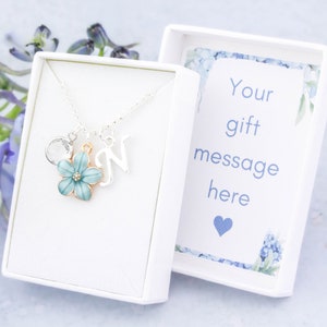 Puede incluir: Un collar de plata con un dije de flor azul, un dije circular de plata y un dije de letra N de plata. El collar está en una caja de regalo blanca con una tarjeta blanca que dice "Su mensaje de regalo aquí" con un corazón azul.