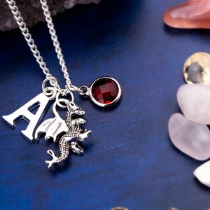 May include: Silver necklace with a silver letter A charm, a silver dragon charm, and a red gemstone charm.