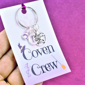 Puede incluir: Un llavero plateado con un dije de cristal morado, un dije de luna creciente, un dije de bruja y un dije de pentagrama. El llavero est&aacute; unido a una tarjeta blanca con el texto "Coven Crew" y un gr&aacute;fico de escoba.