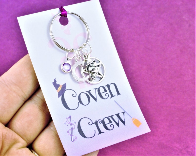 Puede incluir: Un llavero plateado con un dije de cristal morado, un dije de luna creciente, un dije de bruja y un dije de pentagrama. El llavero est&aacute; unido a una tarjeta blanca con el texto "Coven Crew" y un gr&aacute;fico de escoba.