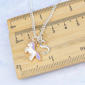 Op de afbeelding: Zilveren ketting met een eenhoornbedel en een letter 'S'-bedel. De ketting ligt op een houten liniaal met inch- en centimetermarkeringen.