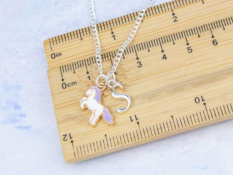 Op de afbeelding: Zilveren ketting met een eenhoornbedel en een letter 'S'-bedel. De ketting ligt op een houten liniaal met inch- en centimetermarkeringen.