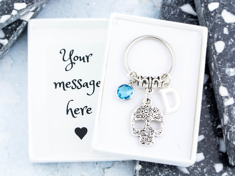 May include: A silver skull keychain with a blue crystal charm, a silver letter D charm, and a silver ring. The keychain is in a white box with the text "Your message here" and a black heart.