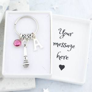 May include: A silver keyring with a microphone charm, a pink crystal, and a letter A charm. The keyring is in a white box with the text "Your message here" and a small heart.