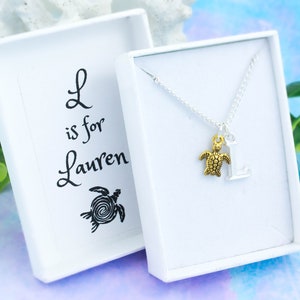 May include: A silver necklace with a gold turtle charm and a silver letter L charm. The necklace is in a white box with the text "L is for Lauren" and a black turtle illustration.
