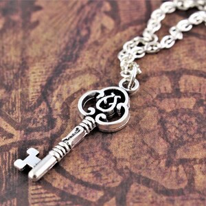 May include: A silver key necklace with a detailed, ornate key pendant. The key has a decorative, swirling design and a small, round bow at the top.
