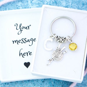 May include: A silver-colored keychain with a yarn and knitting needle charm, a yellow gemstone, and the letter "C". The keychain is presented in a white gift box with the text "Your message here" and a black heart.