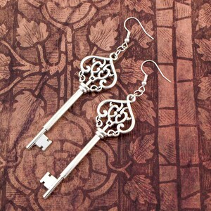 May include: A pair of silver key-shaped earrings with ornate details. The earrings are hanging against a brown patterned background.