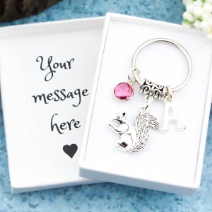 May include: A silver keychain with a squirrel charm, a pink crystal, a silver letter A charm, and a silver ring. The keychain is in a white gift box with the text "Your message here" on the side.