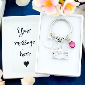 May include: A white gift box containing a silver sewing machine keyring. The keyring features a pink crystal charm and a letter G charm. The box includes a blank card with the text "Your message here" and a black heart.