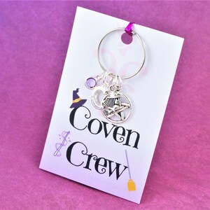 Puede incluir: Una tarjeta blanca con el texto "Coven Crew" y una peque&ntilde;a escoba impresa en ella. Un llavero plateado con una piedra preciosa p&uacute;rpura, un amuleto de luna creciente y un amuleto de pentagrama est&aacute; unido a la tarjeta.